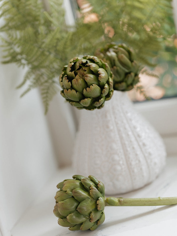 Two green artichokes in a white textured vase with green fern leaves, set on a white surface with a soft blurred background.