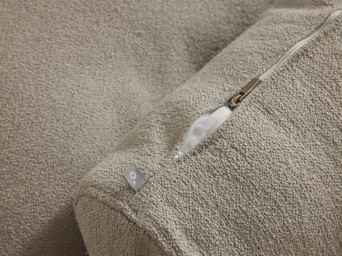 Close-up of a beige fabric cushion with a visible zip and small metal tag on a textured surface.