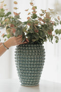 A textured ceramic vase with a blue-green glazed finish, filled with eucalyptus branches, with a person's hand adjusting the foliage.