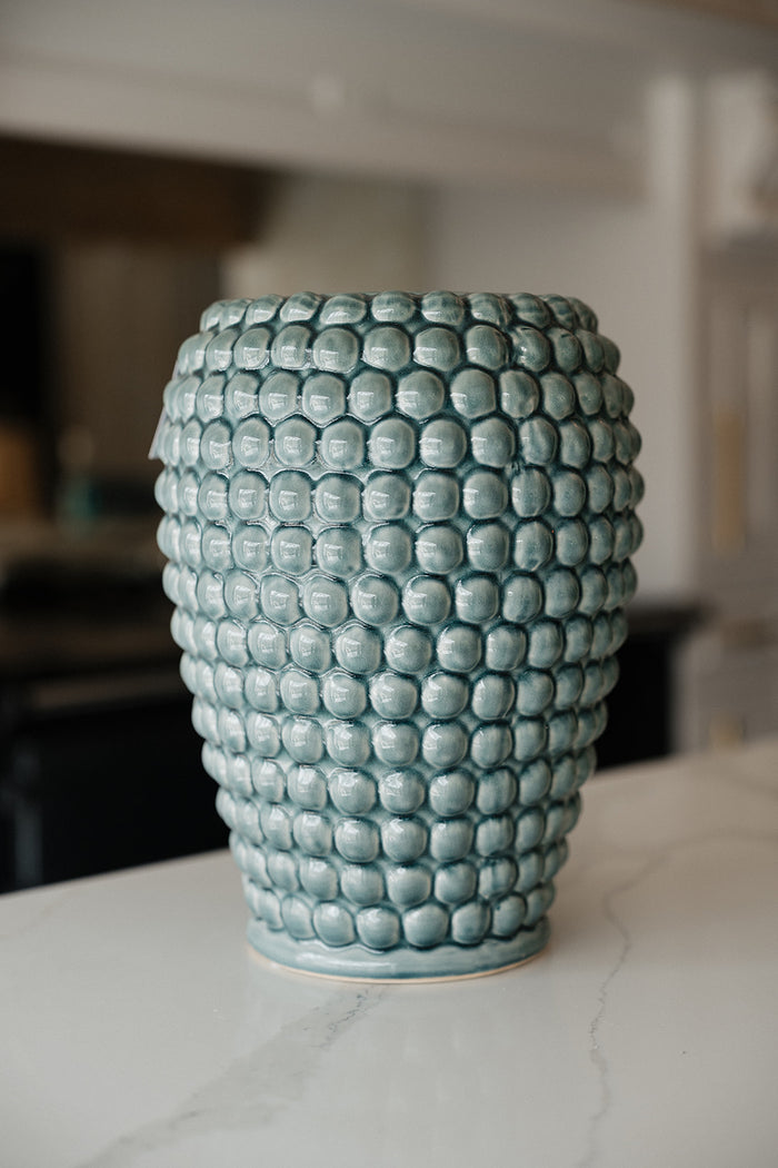 A light blue-green ceramic vase with a textured, raised bubble pattern, sitting on a white marble countertop with a blurred kitchen background.