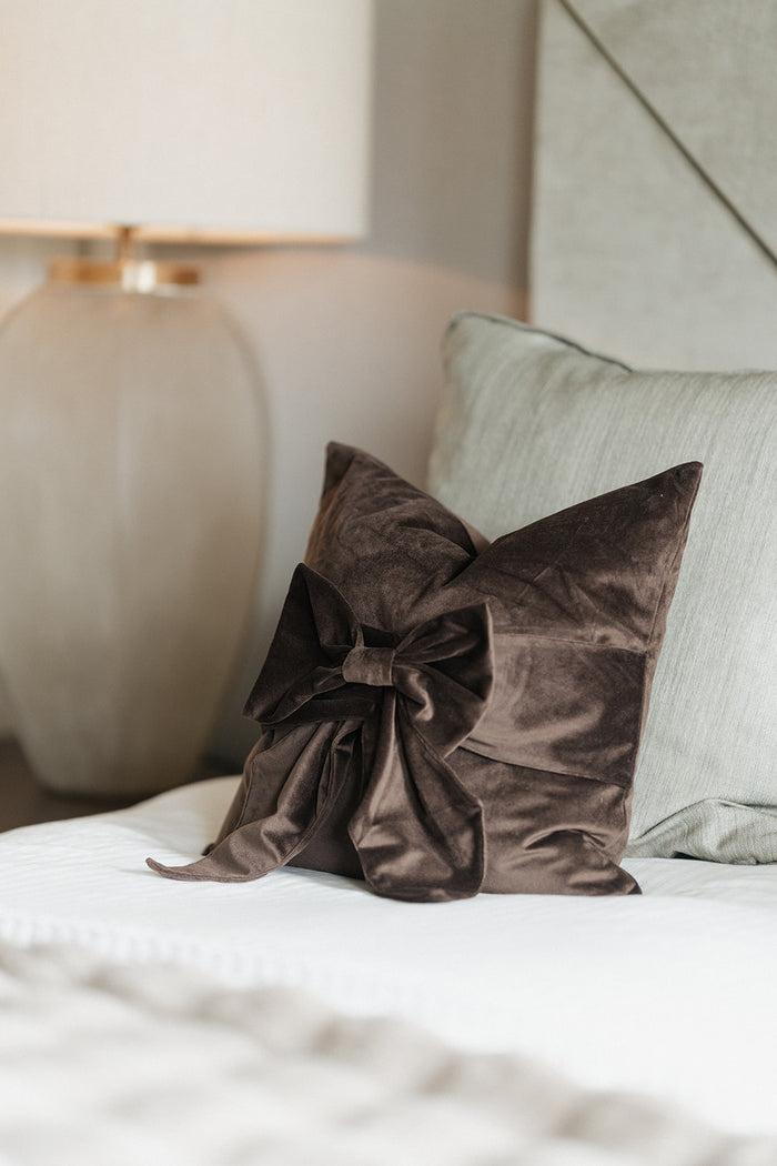A dark brown velvet decorative pillow with a large gathered bow, styled on a white bed next to a light gray pillow and a bedside lamp.
