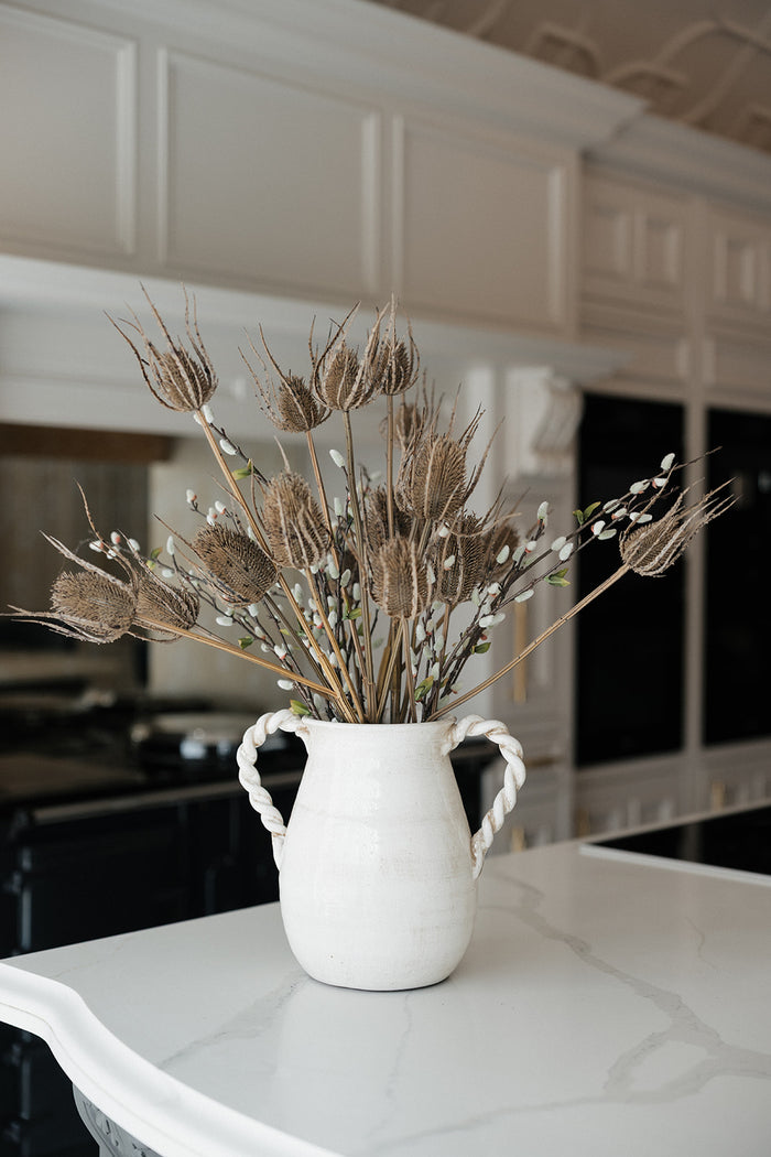 A white ceramic pitcher vase with twisted side handles, filled with dried wheat stalks and small white blossoms, set on a white marble kitchen island with white cabinetry in the background.