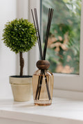 A reed diffuser bottle with amber liquid and dark reeds sits on a white windowsill next to a potted green topiary tree.