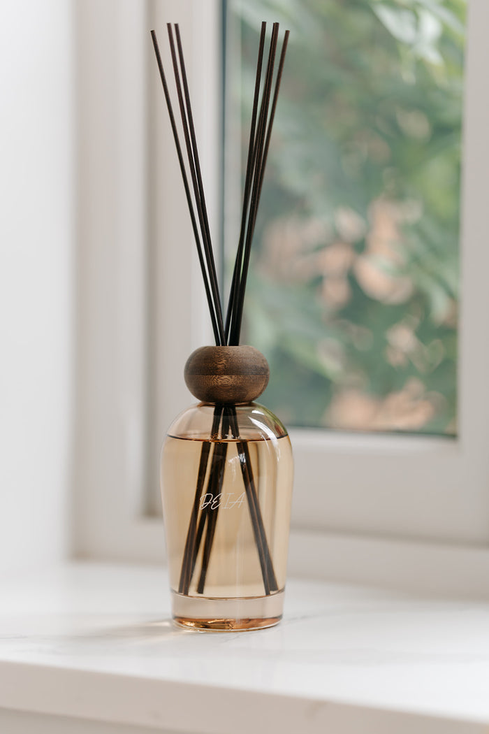 A reed diffuser with a clear glass bottle filled with amber-colored liquid, a round wooden stopper, and several dark brown reeds, placed on a white windowsill with a blurred window view in the background.