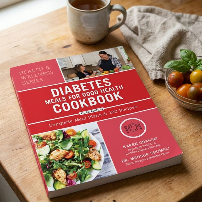 Cookbook titled 'Diabetes Meals for Good Health Cookbook' on a wooden table with a cup of tea and tomatoes.