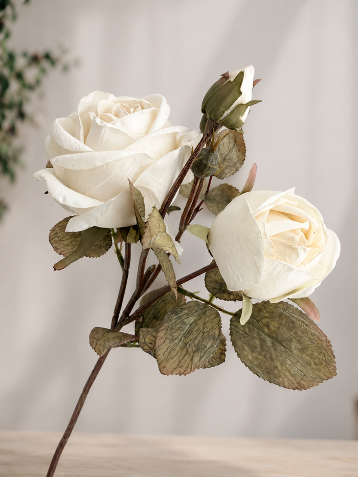 An artificial rose stem featuring two cream-colored roses, one fully bloomed and one bud, with textured olive-green leaves on a light wood surface against a soft beige background.