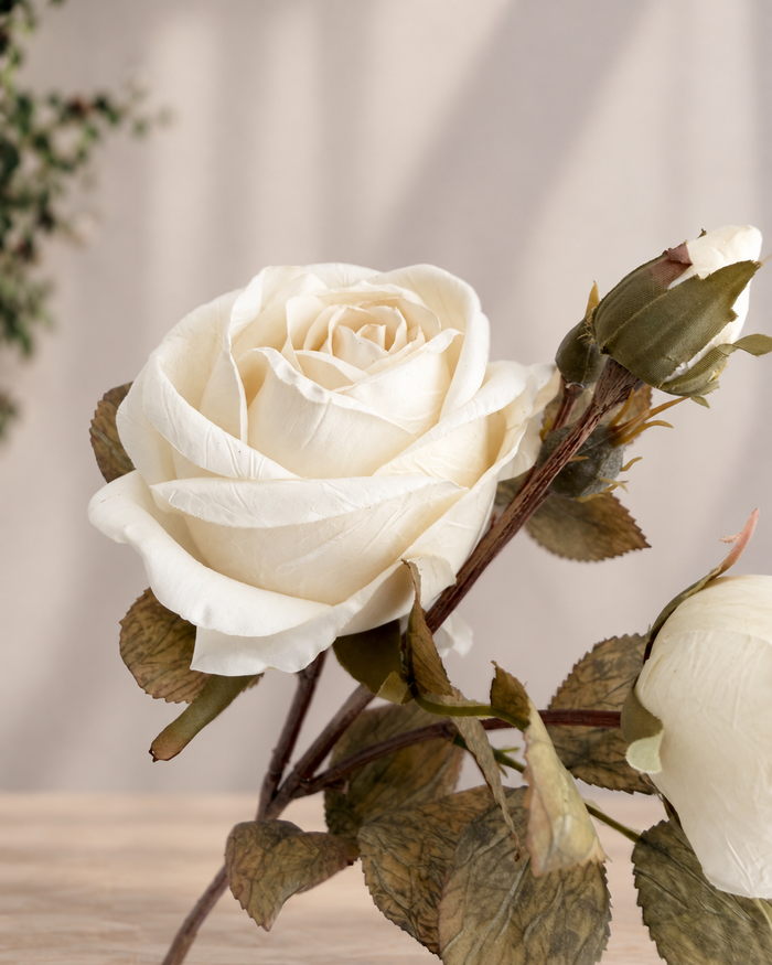 An artificial white rose stem with realistic cream-white petals, brown stems, and textured olive-green leaves, shown against a soft beige background.