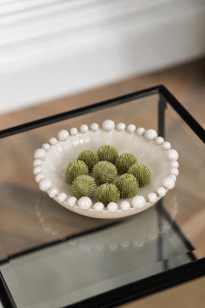 A white ceramic bowl with a beaded border, filled with green decorative pom-pom balls, placed on a glass-top side table.
