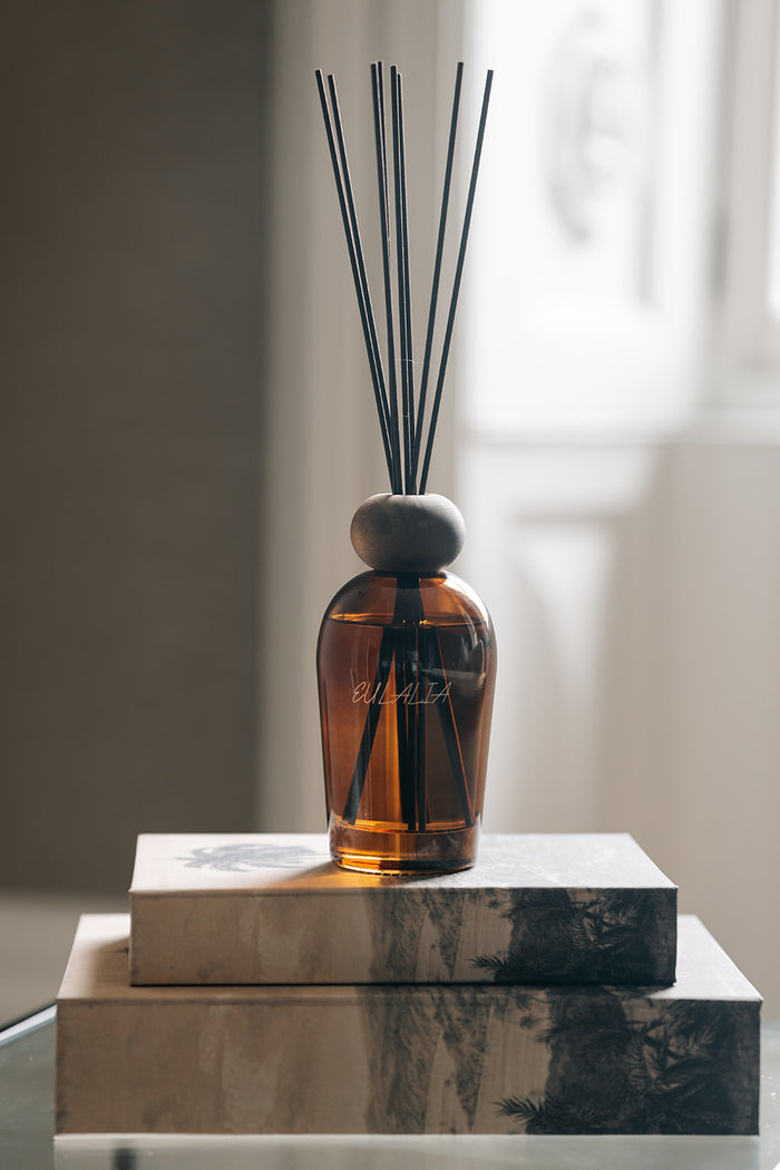 An amber glass reed diffuser bottle with black diffuser sticks and a round gray stopper, displayed on stacked rectangular boxes in a softly lit interior setting.