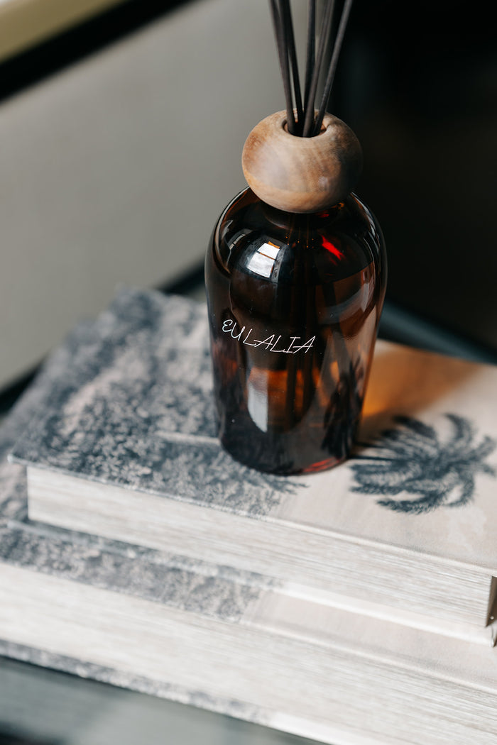 An amber glass reed diffuser bottle with a round wooden top and black reeds, resting on stacked books. The bottle is labeled 'OULALIA'.