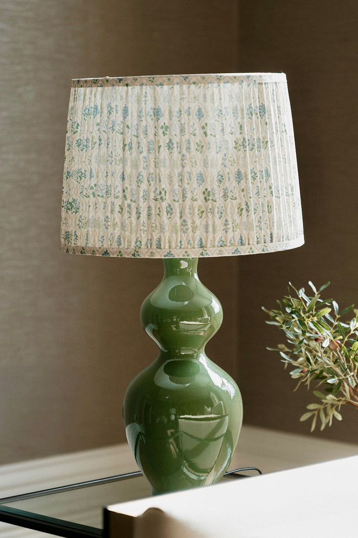 A table lamp with a glossy green ceramic double-bulb base and a pleated fabric shade featuring a light beige and blue-green floral pattern, styled on a tabletop next to a small plant.