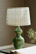 A table lamp with a glossy green ceramic double-bulb base and a pleated fabric shade featuring a light beige and blue-green floral pattern, styled on a tabletop next to a small plant.
