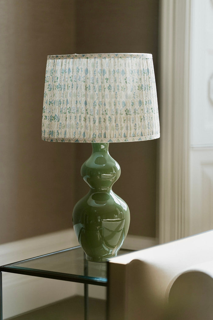A table lamp with a glossy sage green double-bulb vase base and a pleated drum shade featuring a light blue botanical pattern, shown on a glass side table in a softly lit room.