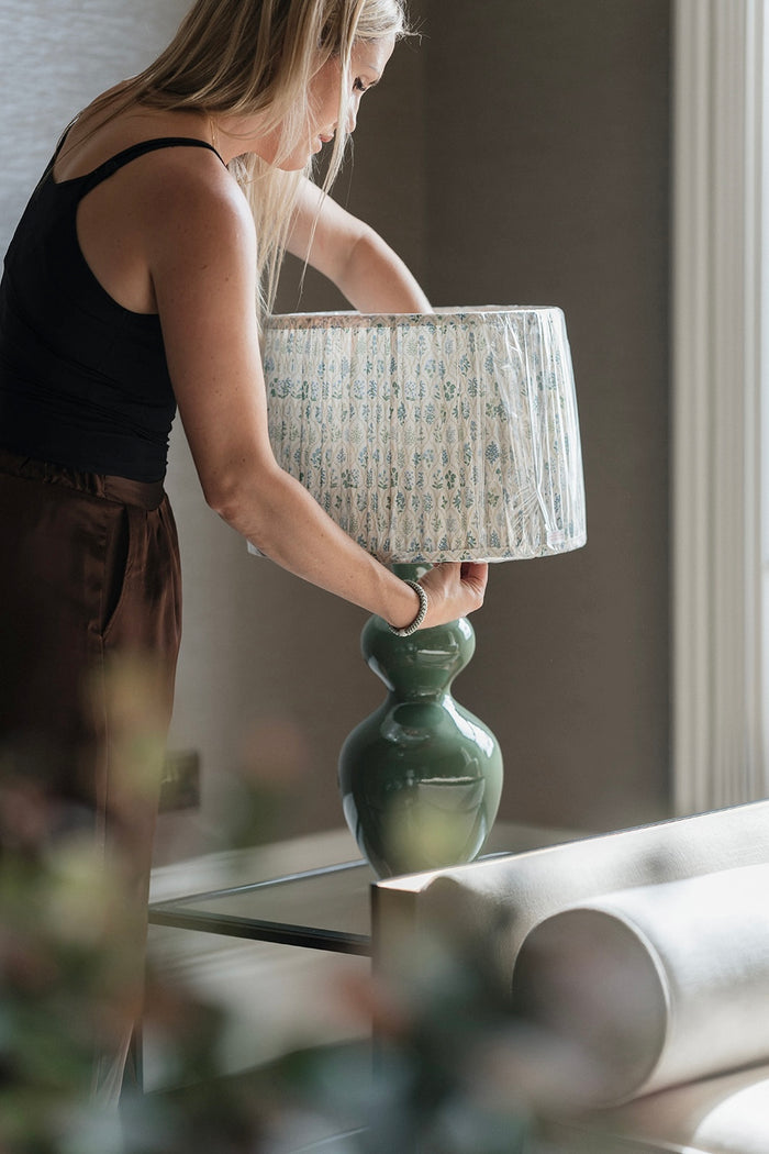 A woman in a black tank top and brown pants adjusts a table lamp with a green ceramic base and a white patterned drum shade in a softly lit indoor setting.