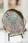 A round decorative serving tray with a metallic gold-toned rim and handles, featuring a vintage floral pattern in blue, pink, and green on a light background, displayed on a small stand.