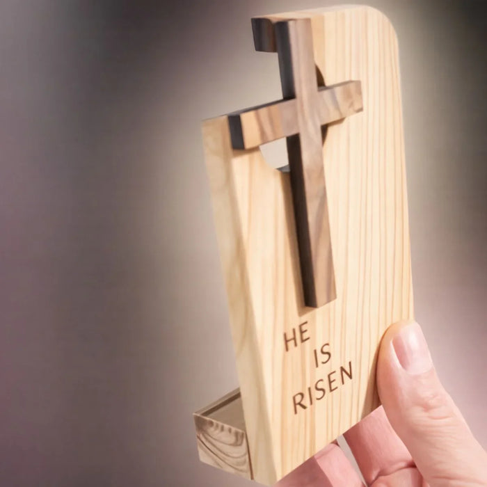 Wooden cross with 'He is Risen' engraving held by a hand against a neutral background
