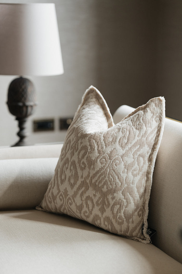 A square decorative throw pillow with a beige and cream damask pattern, resting on a light beige sofa next to a lit table lamp.