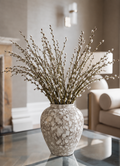 A ceramic vase filled with dried willow branches, set on a glass coffee table in a softly lit living room.
