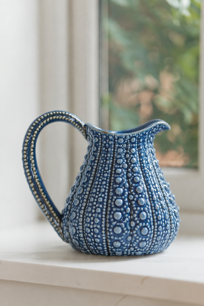 A blue ceramic pitcher with a textured, raised dot pattern and a curved handle, sitting on a white windowsill with a blurred window and greenery in the background.