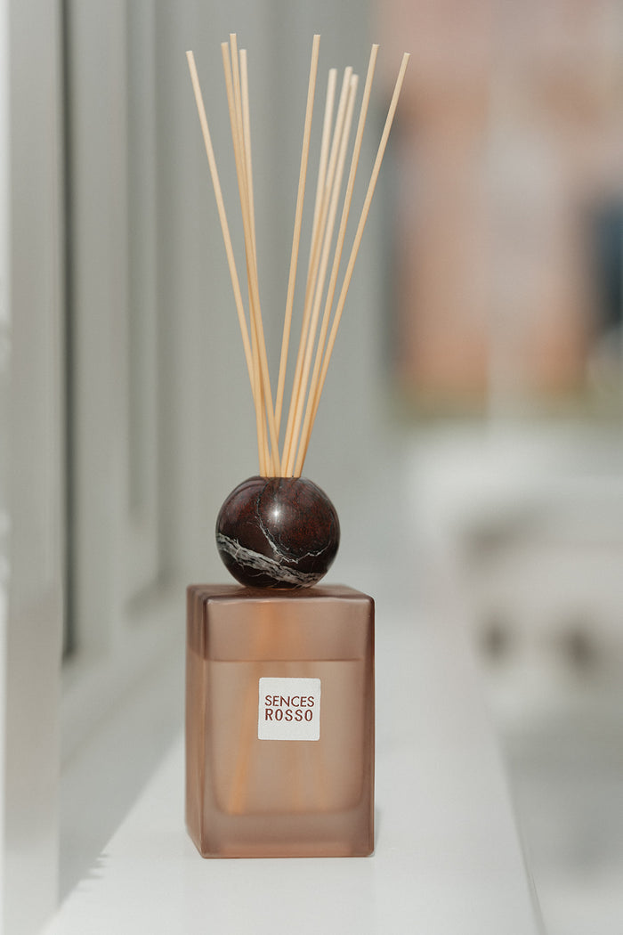 A reed diffuser bottle with a frosted amber-brown glass base, a dark polished stone sphere stopper, and natural reed sticks, shown on a white surface with a softly blurred indoor background. The front label reads 'SENSES ROSSO'.