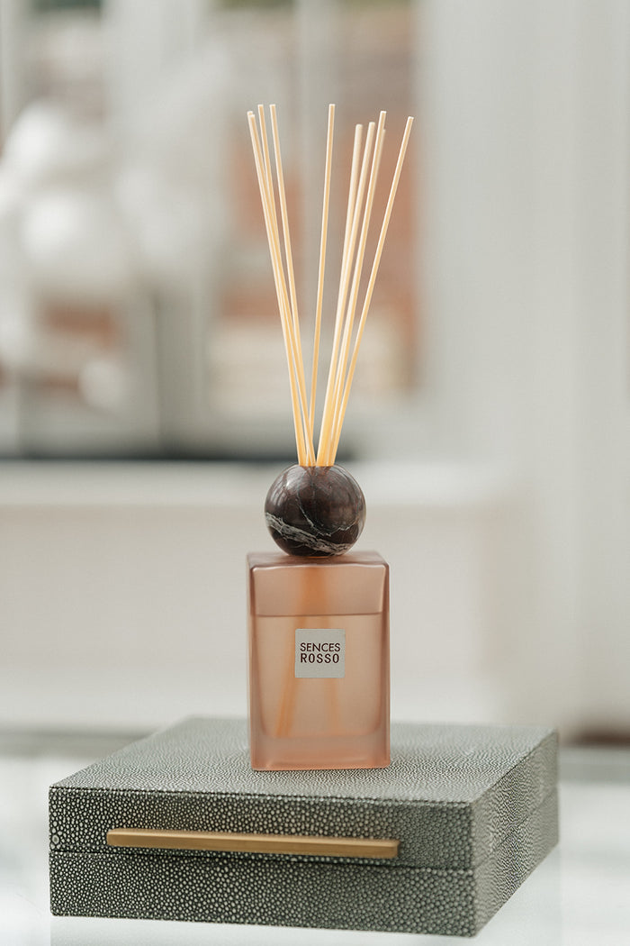 A reed diffuser bottle with a frosted amber-brown glass body, a dark round stopper, and natural reed sticks, displayed on top of a textured gray box with a gold handle.