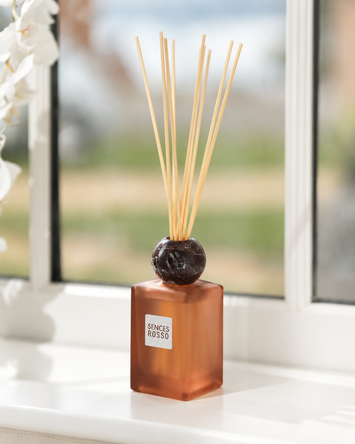 A reed diffuser with a frosted amber-brown glass bottle, a dark round stopper, and natural reed sticks, sitting on a white windowsill with a blurred outdoor background.