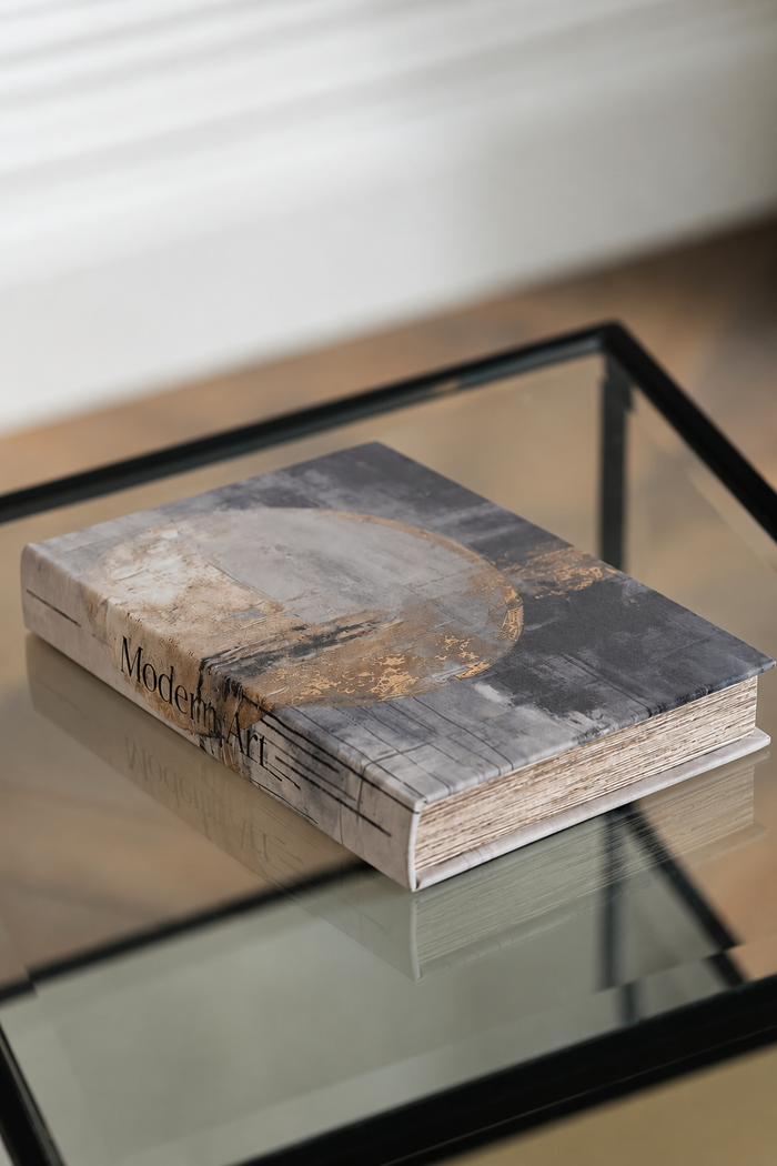 A hardcover art book titled 'Modern Art' resting on a glass-topped table. The book cover features a textured abstract design in gray, black, and gold tones.