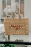 A natural woven straw tote bag with two top handles and the word 'Norfolk' embroidered in red across the front, displayed on a glass table in a softly blurred indoor setting.