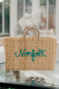 A natural woven straw tote bag with two rolled handles and the word 'Norfolk' embroidered in teal script across the front, shown on a glass table with a softly blurred indoor background.
