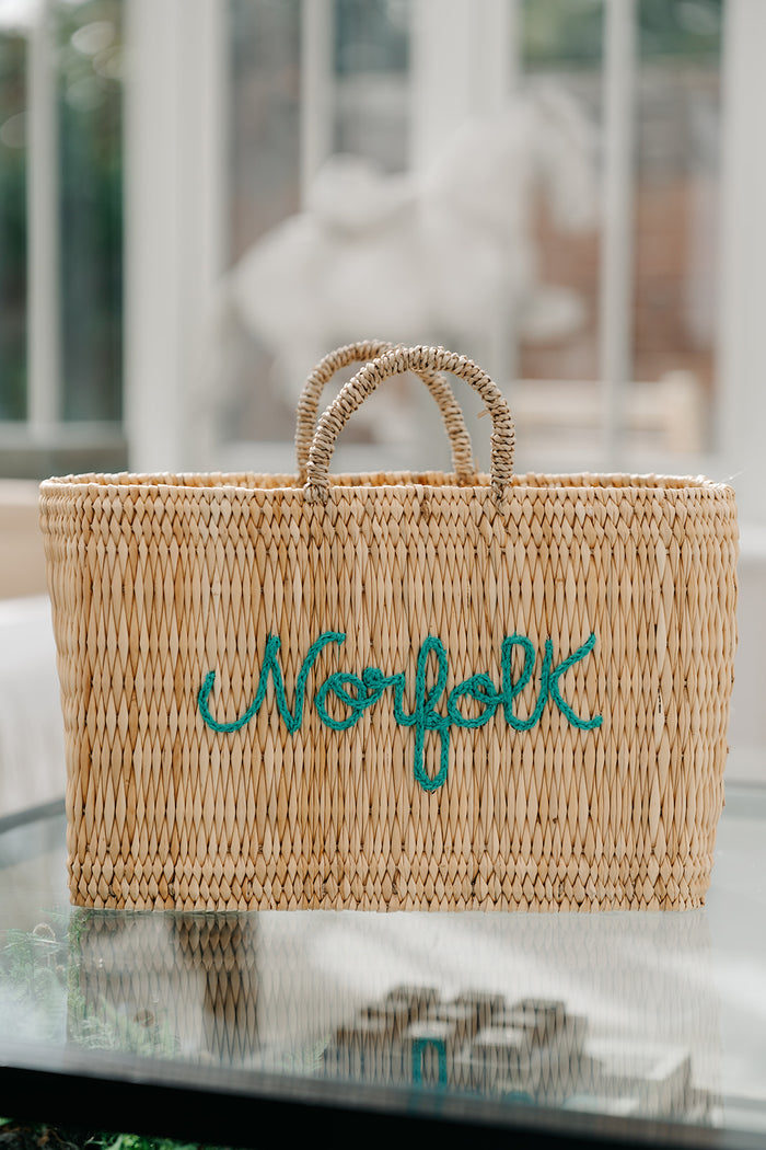 A natural woven straw tote bag with two rolled handles and the word 'Norfolk' embroidered in teal script across the front, shown on a glass table with a softly blurred indoor background.