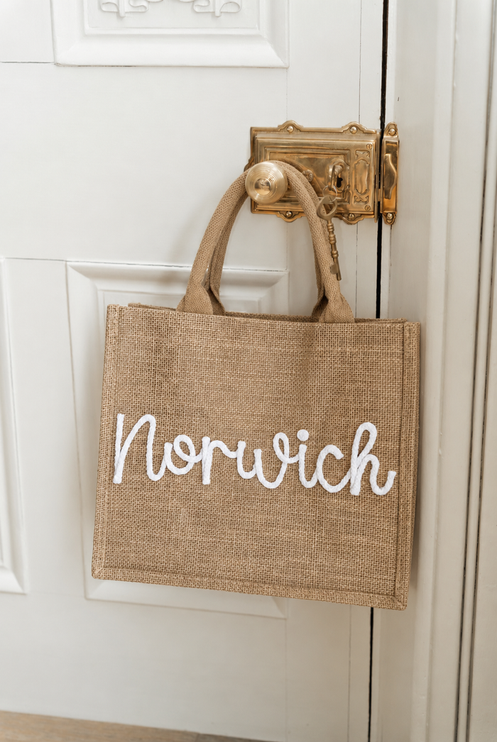 A natural jute tote bag with 'Norwich' written in white script across the front, hanging from a brass doorknob on a white paneled door.