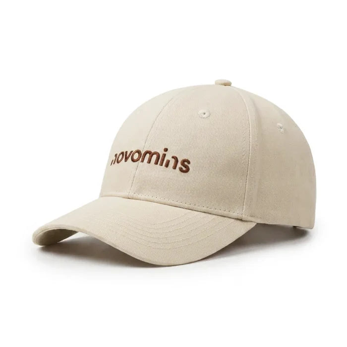 A cream Novomins baseball cap shown at a three-quarter angle on a white background, with the novomins wordmark embroidered in brown across the front panel.