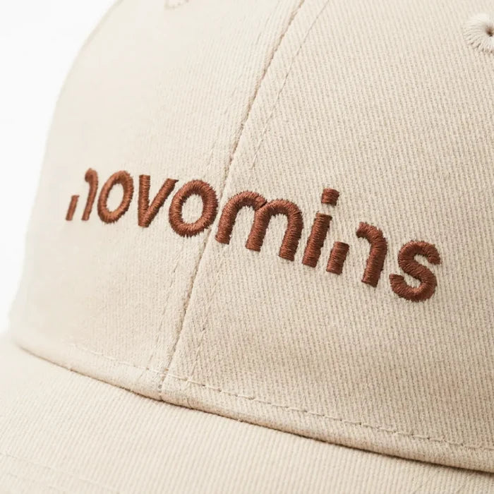 Close-up of the brown embroidered novomins wordmark on the front panel of a cream baseball cap, showing the stitch detail.