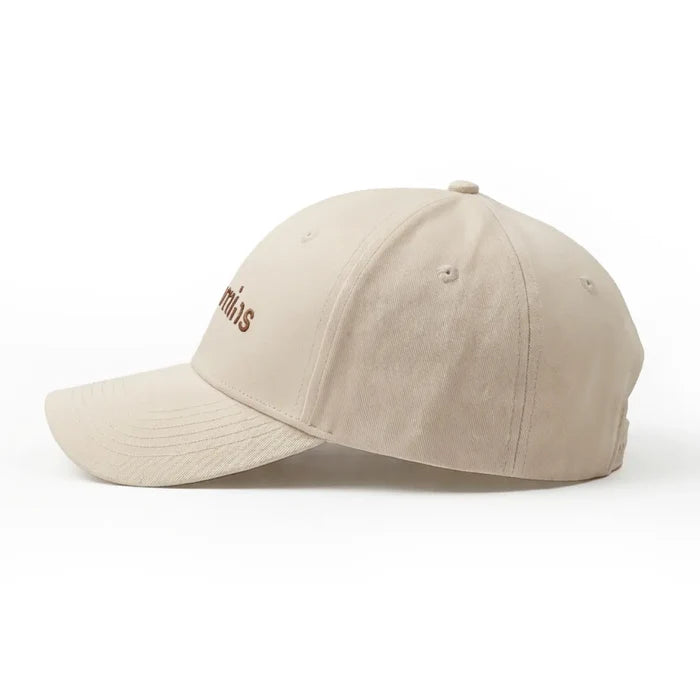 Side profile of a cream Novomins baseball cap on a white background, with the edge of the brown embroidered wordmark visible.
