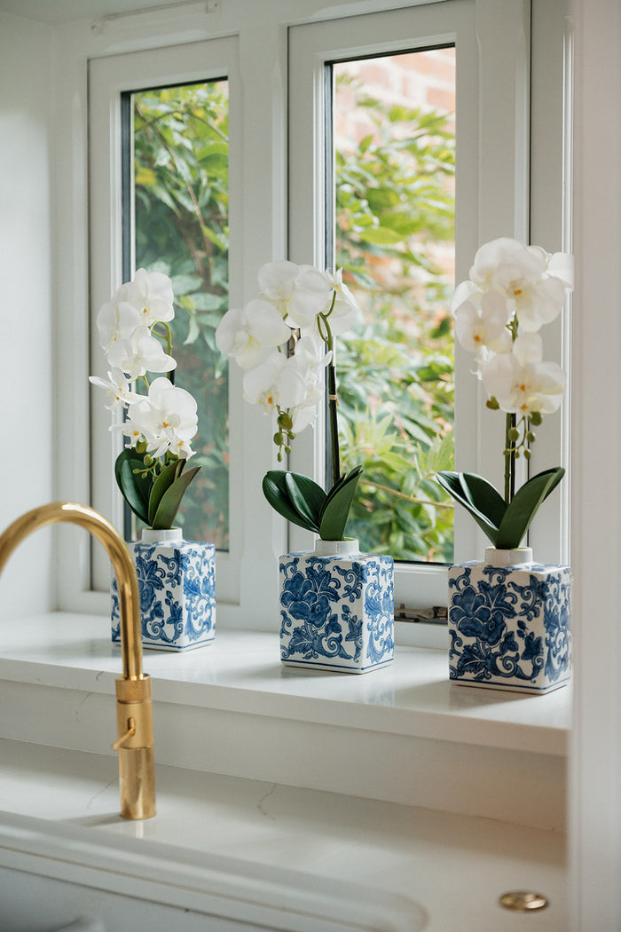 Three white Phalaenopsis orchid plants in blue and white patterned ceramic vases, arranged on a white windowsill with a gold faucet in the foreground and green foliage visible outside the window.