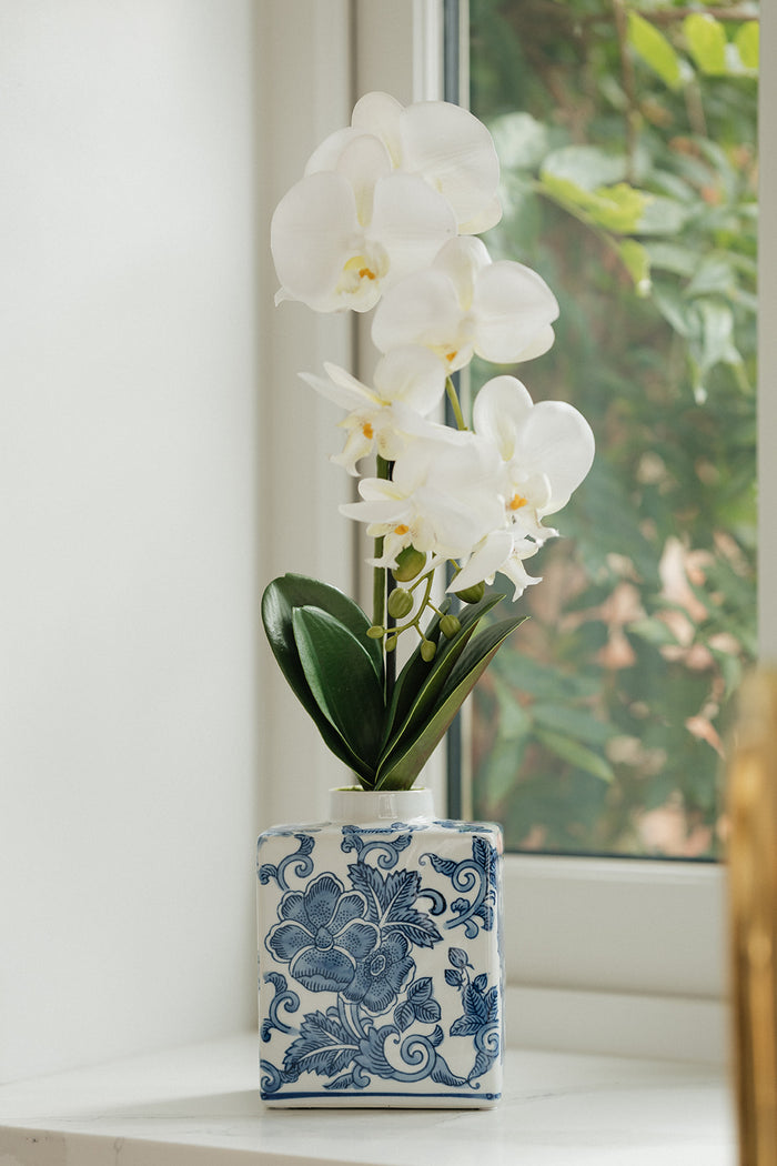 White artificial orchid flowers with green leaves arranged in a blue and white patterned ceramic vase, set on a windowsill with a blurred window and greenery in the background.