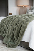 An olive green chunky faux fur throw blanket draped over a bed with white chevron bedding in a softly lit bedroom setting.