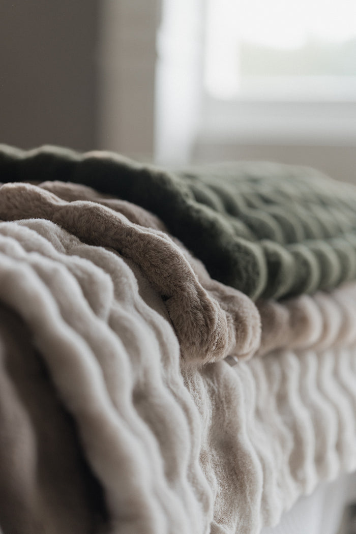 Three textured, ribbed knit blankets in cream, taupe, and olive green stacked on a bed in a softly lit bedroom.