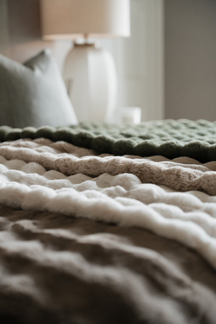A close-up of a cozy textured throw blanket with chunky knit stripes in olive green, cream, taupe, and brown, styled on a bed in a softly lit bedroom.