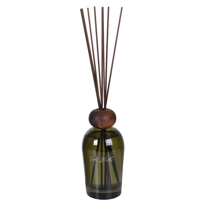 An amber glass reed diffuser bottle with a round wooden stopper and several dark brown reeds, shown on a plain white background.