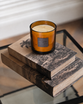 An amber glass jar candle with cream wax sits on a stack of two rustic books on a glass table. The candle label reads 'SENSES CANDLE CO.