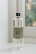 A clear glass bottle of Sences Florist room spray with a black cap and a muted sage green label, standing on a white surface with a softly blurred window background.