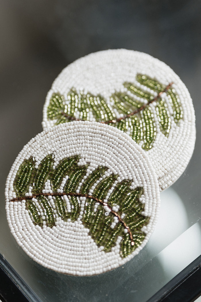 Two round white beaded coasters featuring green beaded fern leaf designs, displayed on a reflective dark surface.