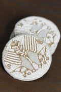 Two round white beaded coasters with gold-toned geometric and floral patterns, stacked on a dark brown wooden surface.